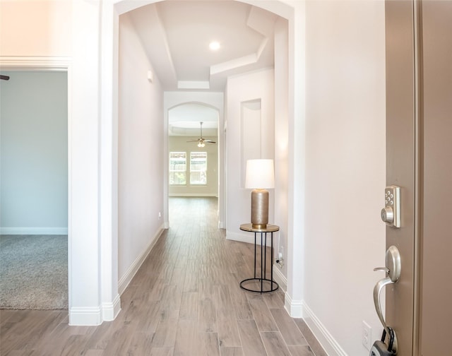 hallway with a raised ceiling, arched walkways, baseboards, and light wood finished floors