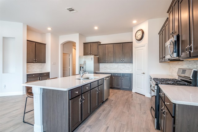 kitchen featuring an island with sink, arched walkways, a sink, light wood finished floors, and stainless steel appliances