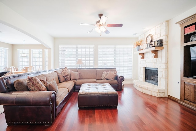 living room featuring dark wood-type flooring, a ceiling fan, arched walkways, and a stone fireplace