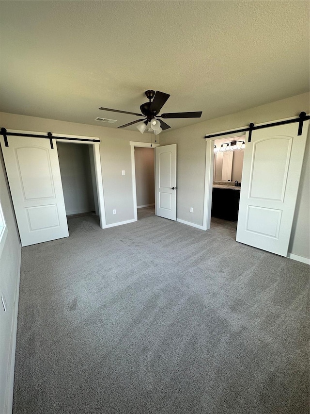 unfurnished bedroom with a textured ceiling, a barn door, carpet flooring, baseboards, and ceiling fan