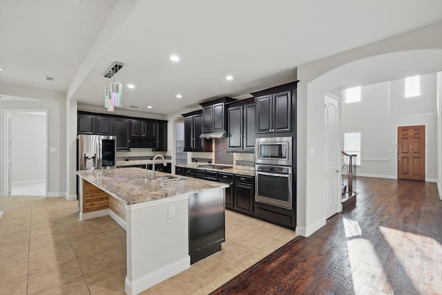 kitchen with light stone countertops, a kitchen island with sink, arched walkways, a sink, and stainless steel appliances