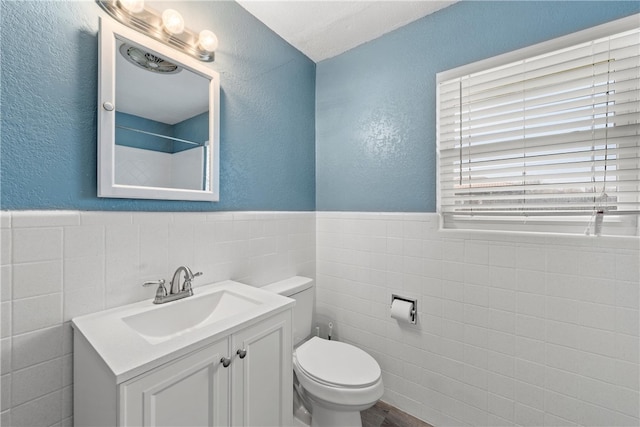 bathroom with toilet, tile walls, wainscoting, vanity, and a textured wall