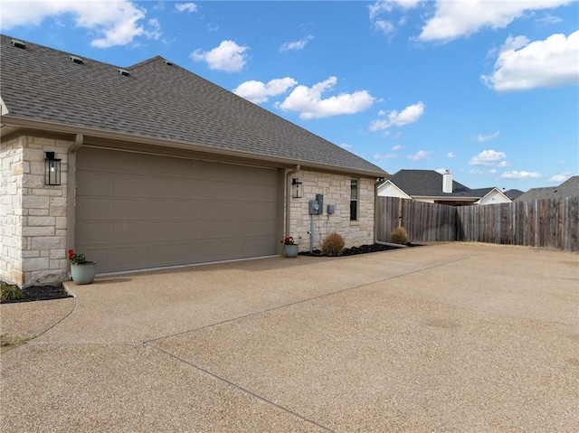 view of side of home featuring a garage, roof with shingles, fence, stone siding, and driveway