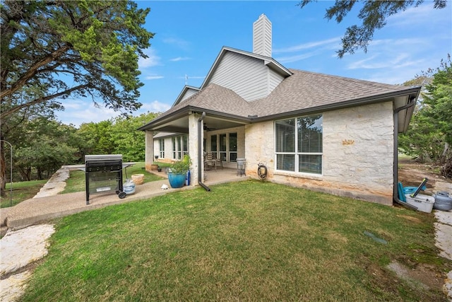 back of house with a shingled roof, a lawn, a chimney, a patio area, and stone siding