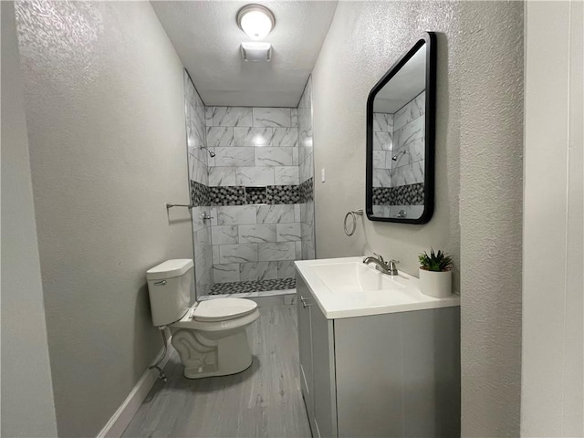 bathroom with toilet, wood finished floors, tiled shower, a textured wall, and vanity
