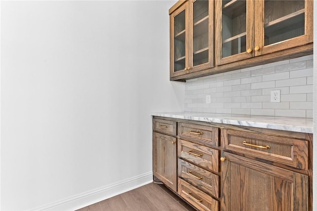 kitchen with backsplash, glass insert cabinets, dark wood-type flooring, baseboards, and brown cabinetry