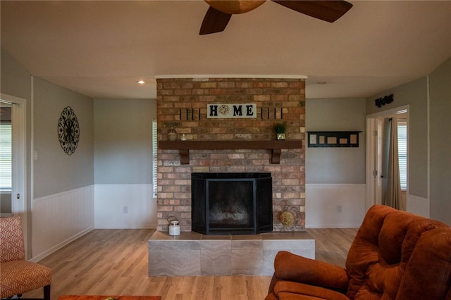 living room featuring a wainscoted wall, a brick fireplace, ceiling fan, and wood finished floors