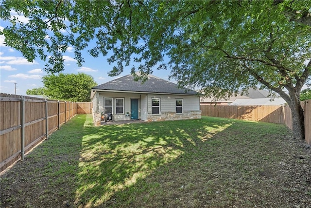 back of property with a fenced backyard, stone siding, a yard, and roof with shingles