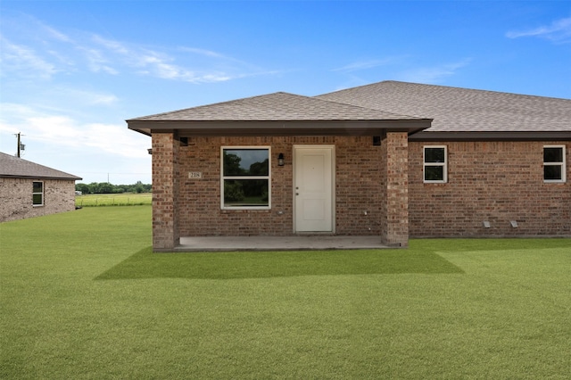 back of property featuring brick siding, a shingled roof, a lawn, and a patio