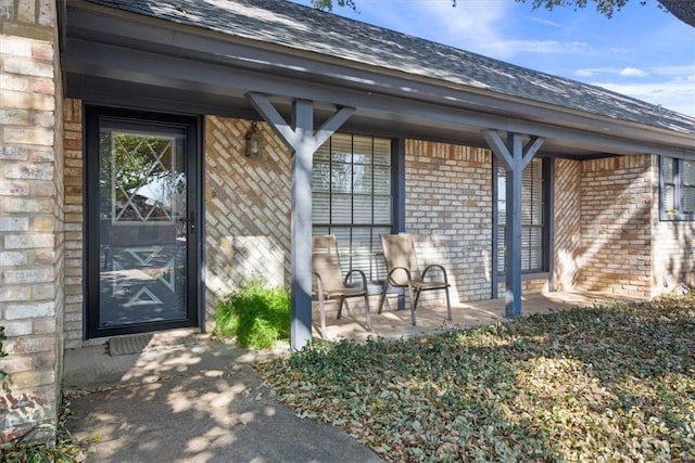 view of exterior entry with brick siding, a porch, and roof with shingles