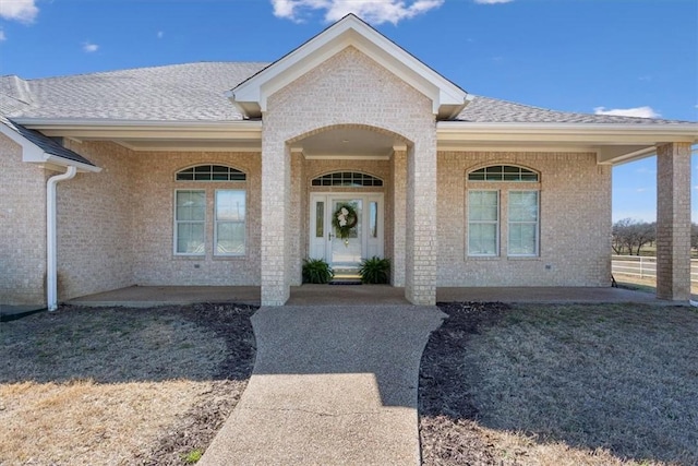 view of exterior entry with brick siding, roof with shingles, and covered porch