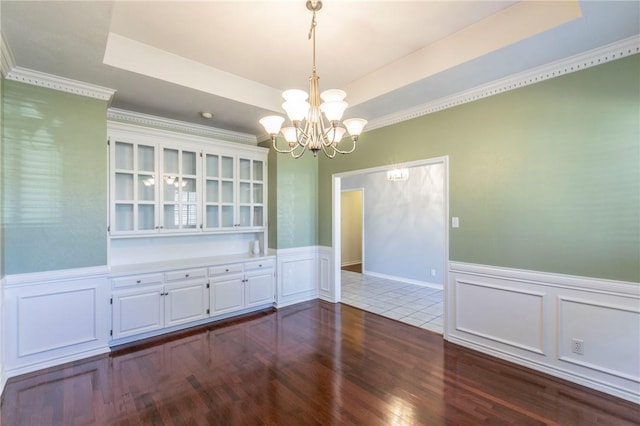unfurnished dining area with wainscoting, an inviting chandelier, dark wood-style floors, a decorative wall, and a raised ceiling