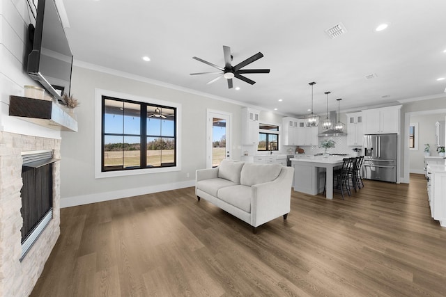 living room with visible vents, ceiling fan, ornamental molding, a stone fireplace, and dark wood-style flooring