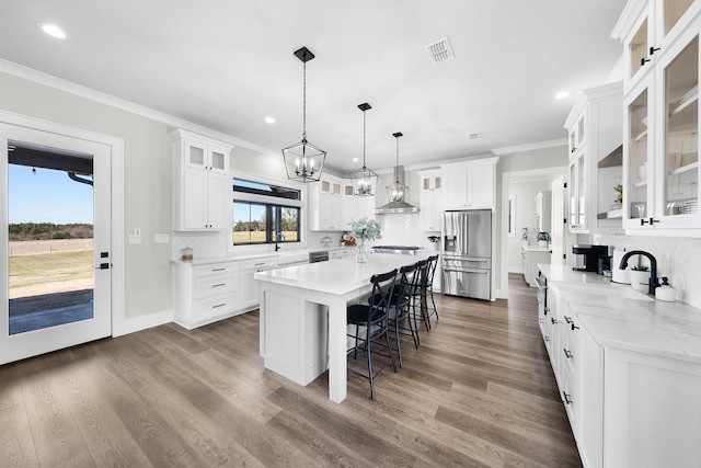 kitchen featuring a center island, a kitchen bar, stainless steel refrigerator with ice dispenser, white cabinets, and wall chimney exhaust hood