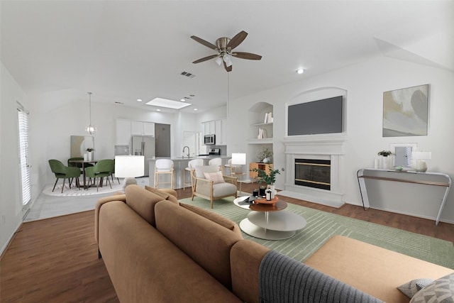 living room with a ceiling fan, dark wood-style floors, a glass covered fireplace, vaulted ceiling, and built in shelves