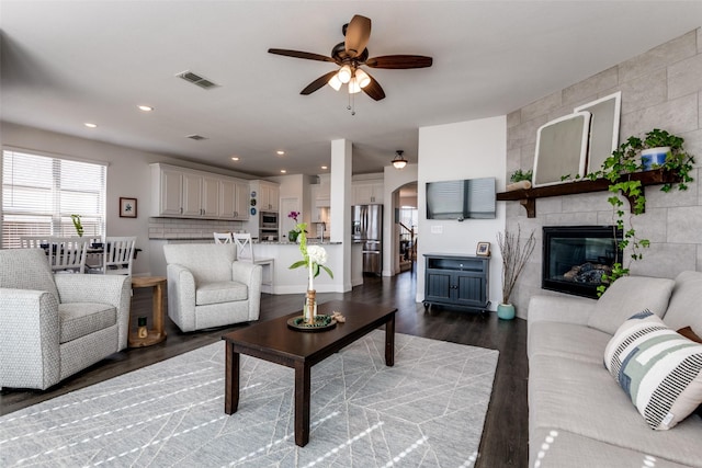 living room with a ceiling fan, arched walkways, a tiled fireplace, dark wood-style flooring, and visible vents