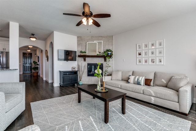 living area featuring arched walkways, dark wood-style flooring, a tiled fireplace, baseboards, and a ceiling fan