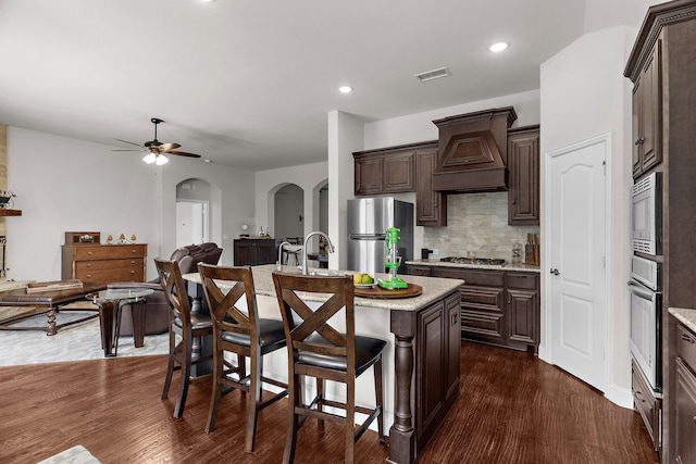 1229 Bridgeport Dr, Anna, TX 75409 - kitchen featuring stainless steel appliances, custom range hood, arched walkways, dark brown cabinets, and a kitchen bar