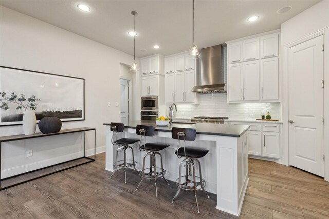 kitchen featuring white cabinets, stainless steel appliances, wood finished floors, wall chimney exhaust hood, and a kitchen bar