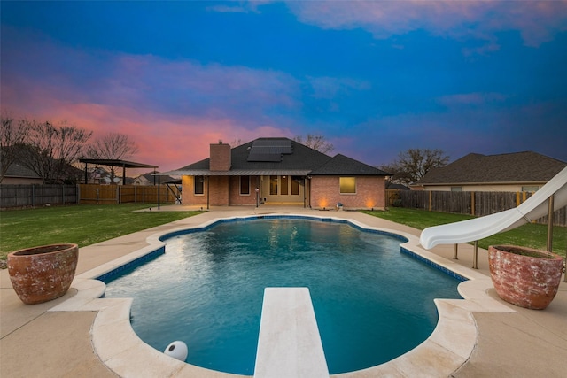 pool at dusk featuring a lawn, a fenced backyard, a water slide, and a patio