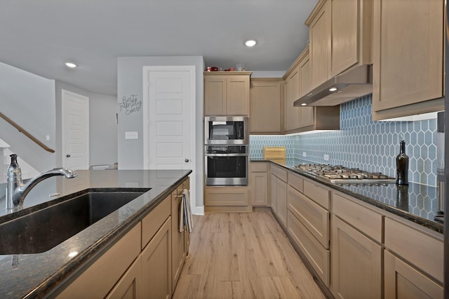 kitchen with dark stone counters, light wood-style floors, under cabinet range hood, appliances with stainless steel finishes, and a sink