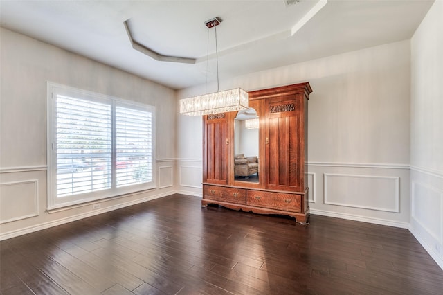 unfurnished dining area featuring dark wood-style floors, a tray ceiling, a decorative wall, wainscoting, and a chandelier