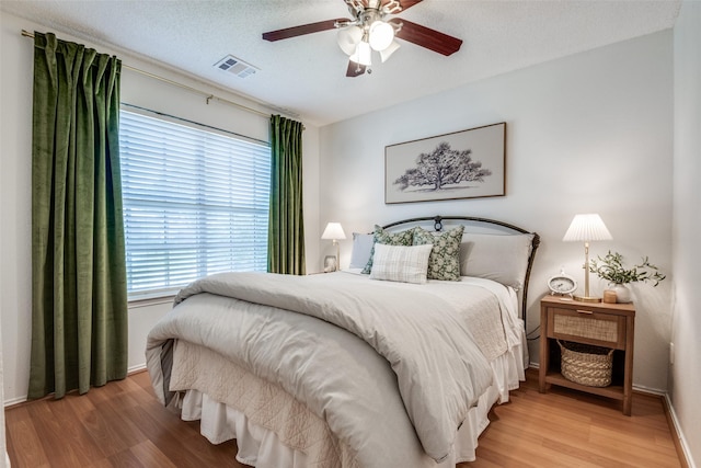 bedroom with a textured ceiling, visible vents, baseboards, light wood finished floors, and a ceiling fan
