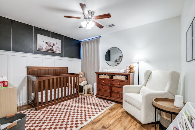 bedroom with visible vents, a crib, light wood finished floors, a decorative wall, and a ceiling fan