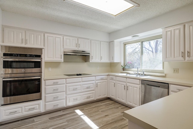 kitchen with white cabinets, appliances with stainless steel finishes, under cabinet range hood, a sink, and light wood finished floors