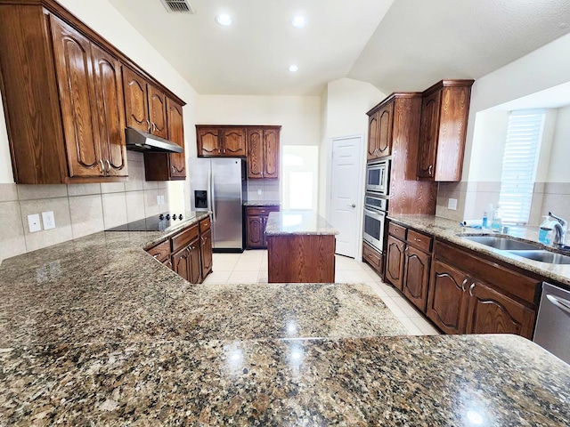 kitchen with under cabinet range hood, stainless steel appliances, a sink, dark stone counters, and decorative backsplash