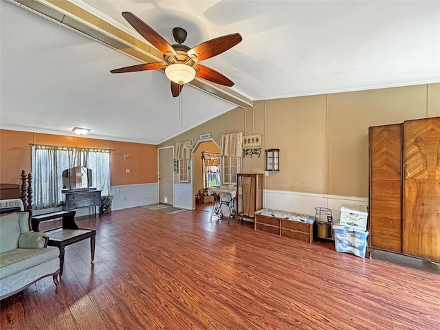 living area with a ceiling fan, vaulted ceiling with beams, wood finished floors, wainscoting, and a decorative wall