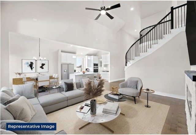 living area featuring ceiling fan with notable chandelier, wood finished floors, stairway, a high ceiling, and baseboards