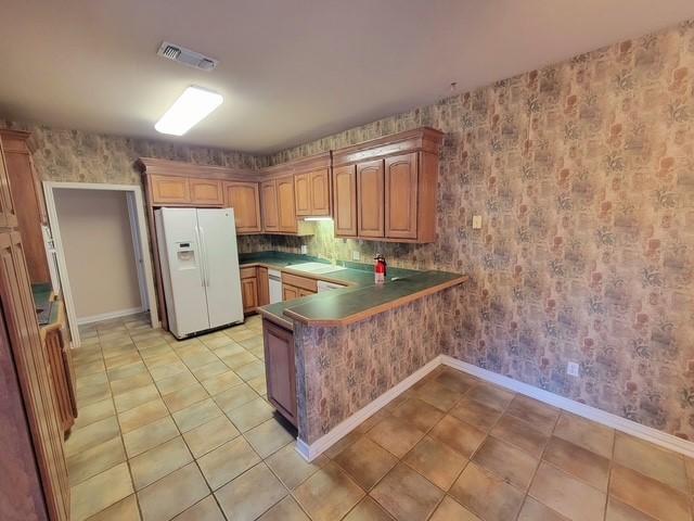 kitchen with wallpapered walls, light tile patterned flooring, visible vents, a peninsula, and white fridge with ice dispenser