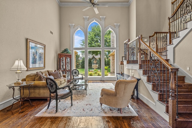 living area featuring a towering ceiling, hardwood / wood-style floors, ornamental molding, ceiling fan, and stairs