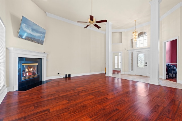 unfurnished living room featuring ornate columns, ornamental molding, wood finished floors, a tiled fireplace, and ceiling fan with notable chandelier