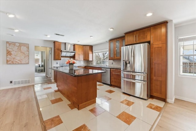 kitchen featuring appliances with stainless steel finishes, wall chimney exhaust hood, a center island, tasteful backsplash, and brown cabinetry