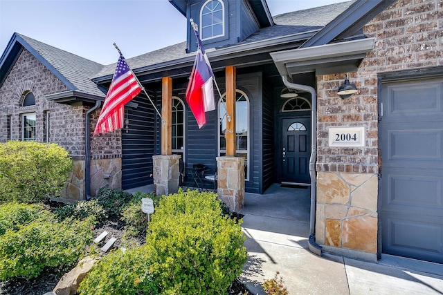 entrance to property with stone siding, covered porch, an attached garage, and roof with shingles