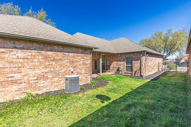 back of house featuring central air condition unit, fence, brick siding, a lawn, and a shingled roof