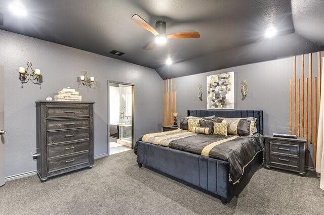 carpeted bedroom featuring ceiling fan with notable chandelier, vaulted ceiling, visible vents, and ensuite bathroom