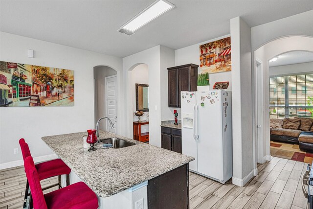 kitchen featuring a sink, white refrigerator with ice dispenser, dark brown cabinetry, arched walkways, and wood finish floors