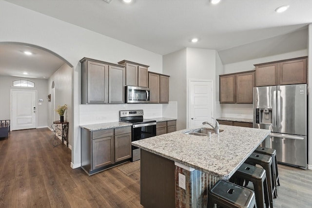 kitchen with a sink, a center island with sink, arched walkways, light stone countertops, and appliances with stainless steel finishes