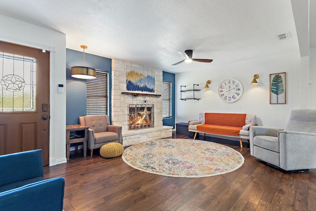 living room with a textured ceiling, visible vents, a ceiling fan, a stone fireplace, and wood finished floors