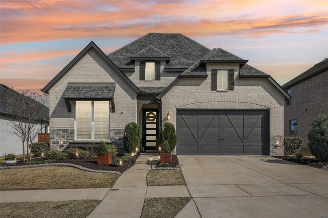 french country inspired facade with roof with shingles, an attached garage, stone siding, driveway, and brick siding