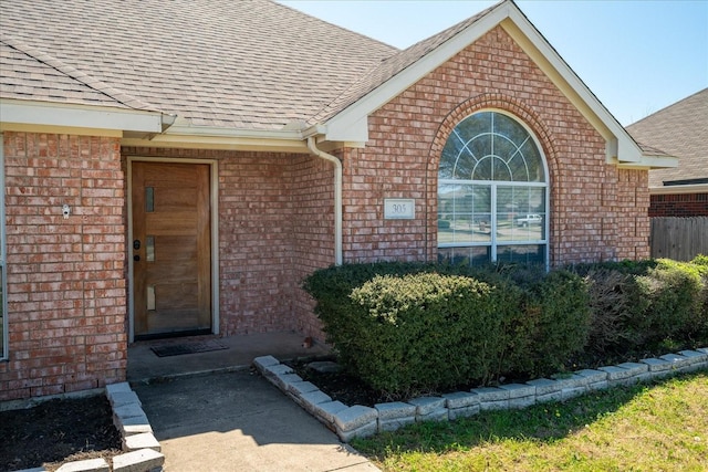 entrance to property featuring a shingled roof, fence, and brick siding
