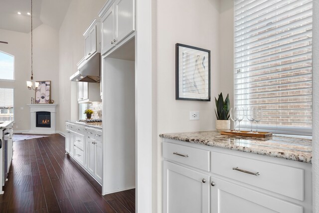 15664 Wild Cherry Ln, Fort Worth, TX 76262 - kitchen with under cabinet range hood, stainless steel gas stovetop, a lit fireplace, white cabinetry, and light stone counters