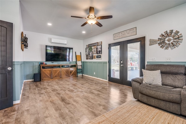 living area with french doors, a ceiling fan, a wall unit AC, wood finished floors, and wainscoting