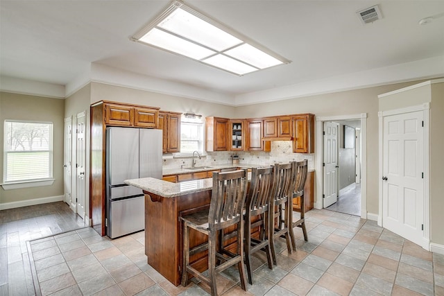 kitchen with a sink, freestanding refrigerator, light stone countertops, a kitchen breakfast bar, and brown cabinets