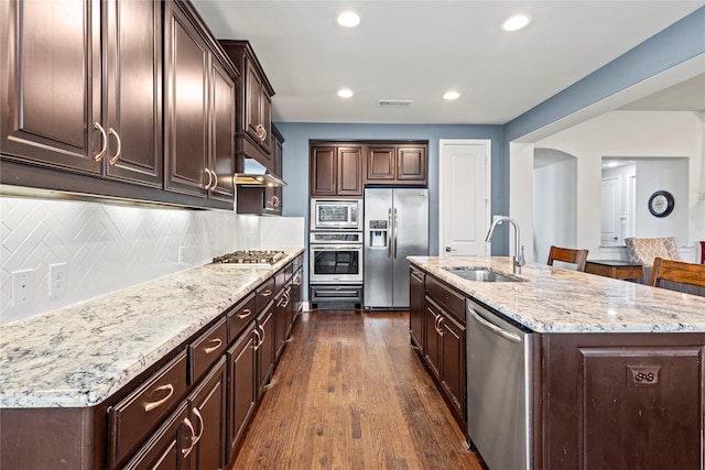 1630 Old Oak Ct, Prosper, TX 75078 - kitchen featuring appliances with stainless steel finishes, dark brown cabinets, a kitchen island with sink, dark wood-type flooring, and light stone countertops