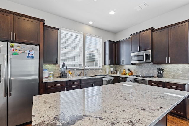 kitchen with stainless steel appliances, a sink, dark brown cabinets, light stone countertops, and tasteful backsplash