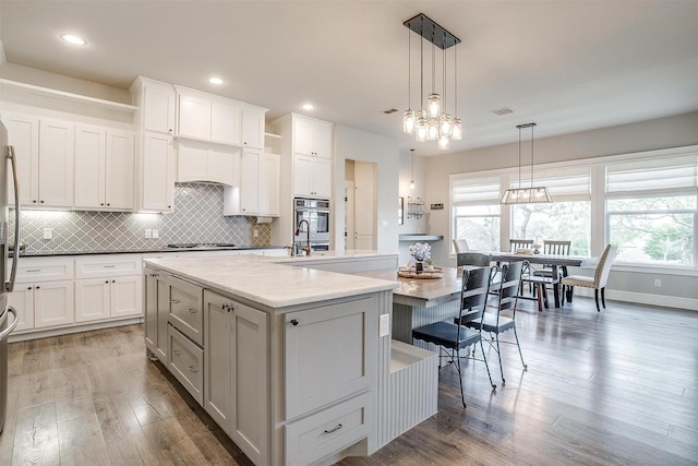 kitchen featuring tasteful backsplash, wood finished floors, hanging light fixtures, a center island with sink, and white cabinetry